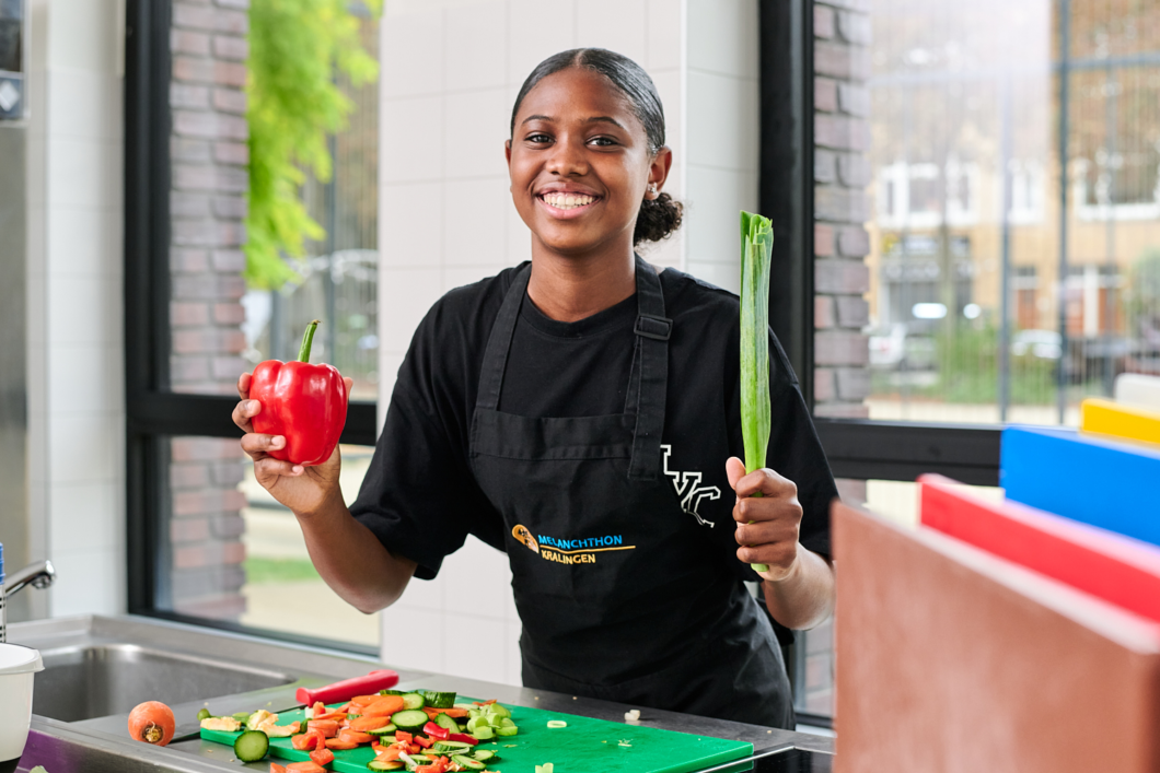 Leerling van Melanchthon Kralingen, staand in de keuken. In haar handen heeft zij een paprika en een prei. Voor haar liggen gesneden groenten op een snijplank.