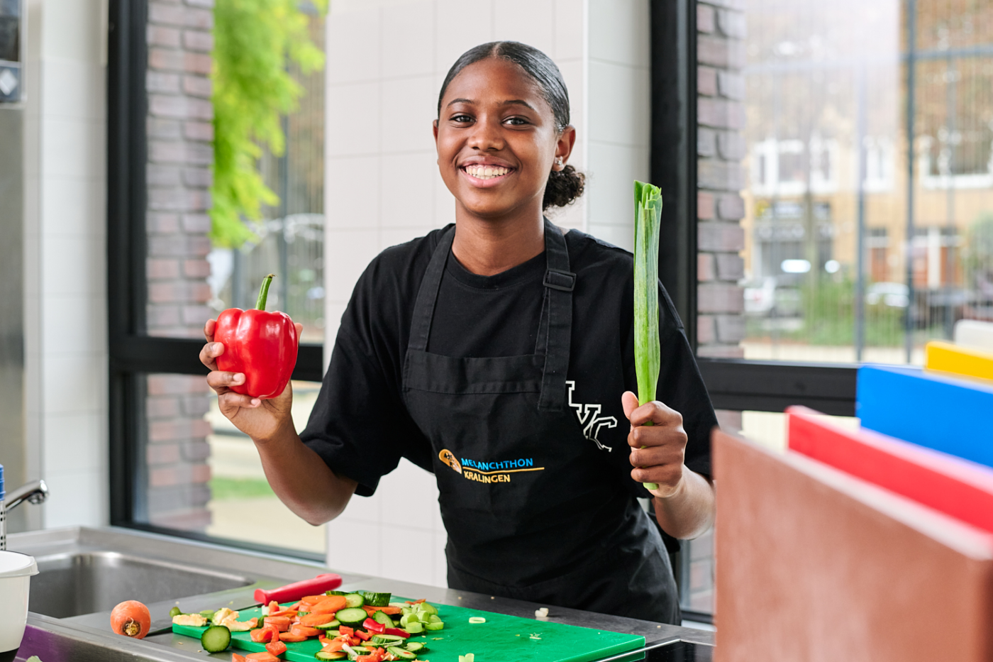 Leerling van Melanchthon Kralingen, staand in de keuken. In haar handen heeft zij een paprika en een prei. Voor haar liggen gesneden groenten op een snijplank.